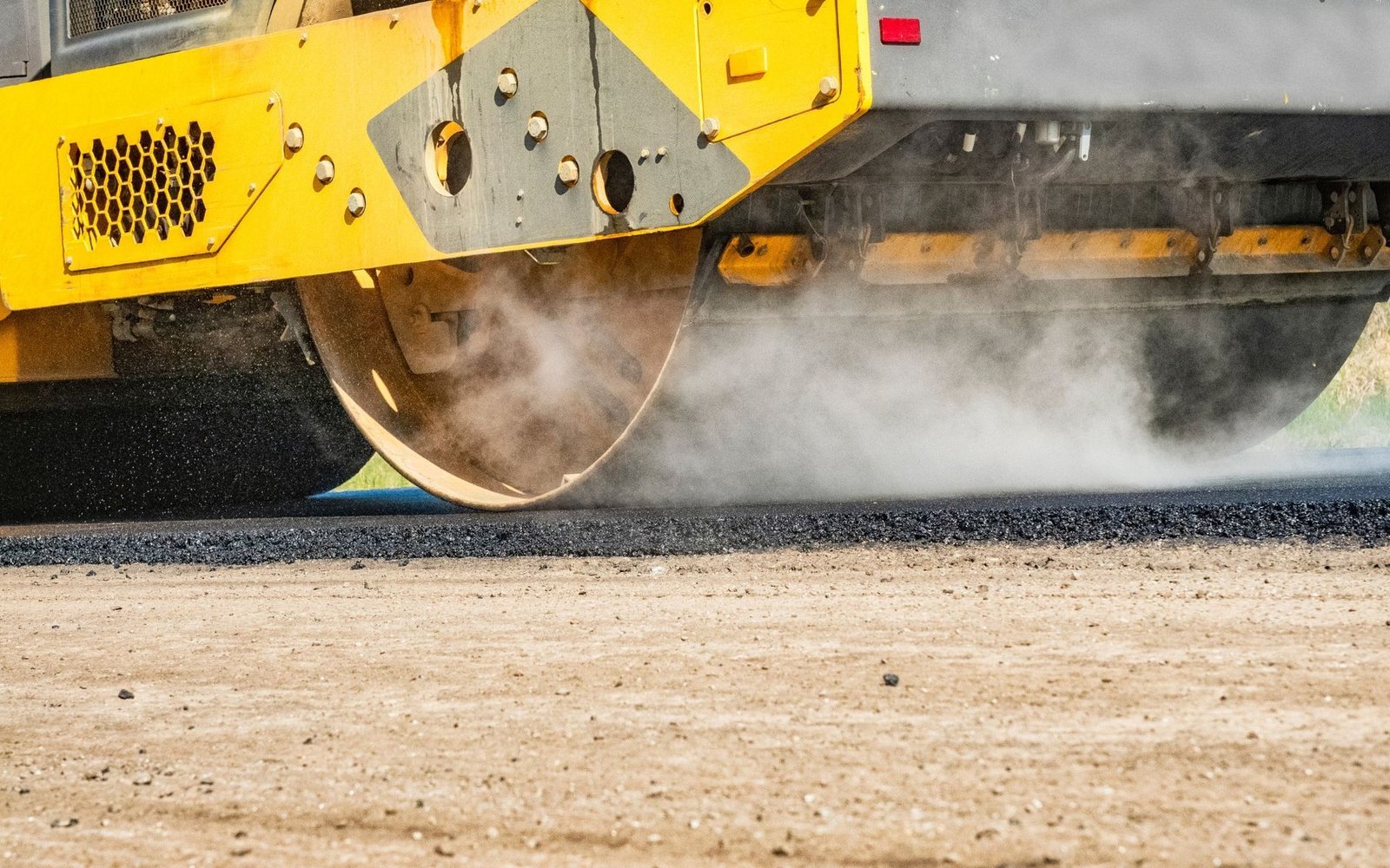 A close-up of a yellow steamroller from an asphalt contractor compacting asphalt on a road in Jensen Beach. The roller is partially visible, with steam rising from the hot asphalt. The dust-covered lower part of the heavy machine highlights ongoing roadwork progress.