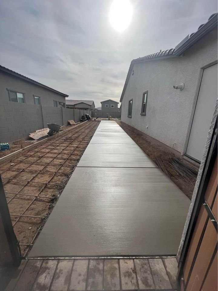 A newly poured concrete pathway stretches between two houses under a cloudy sky. The path, crafted by expert paving services, is smooth and bordered by dirt and construction materials. A distant house is visible at the end of the path.